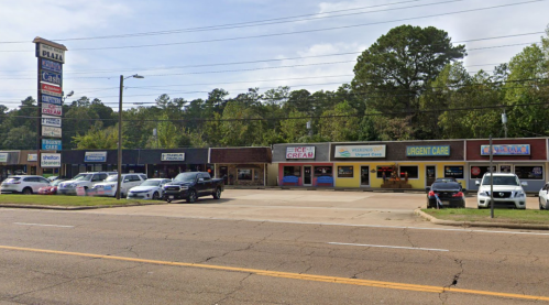 A row of businesses along a street, including an ice cream shop, pharmacy, and urgent care, with parked cars in front.