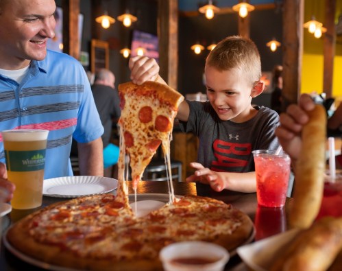 A boy excitedly pulls a slice of pepperoni pizza from a large pizza, while adults enjoy drinks at a lively restaurant.