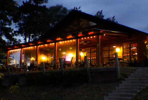 A cozy wooden restaurant illuminated by warm lights, surrounded by trees, with outdoor seating visible.