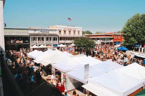 A bustling outdoor market with white tents, crowds of people, and buildings in the background under a clear blue sky.