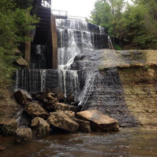 A cascading waterfall flows over rocky terrain, surrounded by lush greenery and a serene atmosphere.