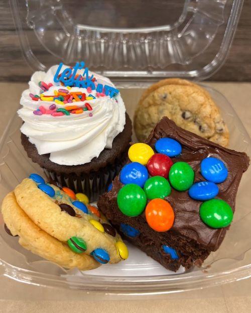 A clear container holds a chocolate cupcake with sprinkles, a cookie, and a brownie topped with colorful candies.
