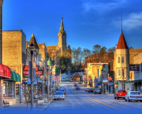 A charming small town street lined with shops, featuring a church steeple in the background under a clear blue sky.