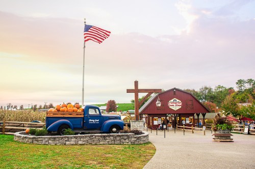 A vintage blue truck with pumpkins parked in front of a barn, with an American flag waving in the background.