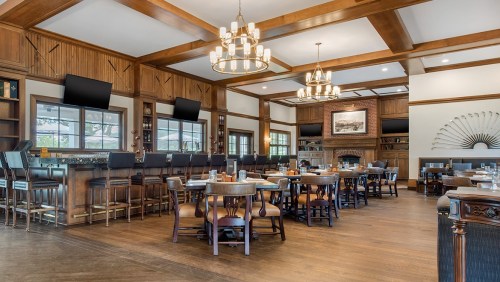 A spacious restaurant interior with wooden beams, tables, and a bar area, featuring chandeliers and large windows.