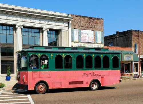 A pink and green trolley bus labeled "The Villages" parked on a street near historic buildings.