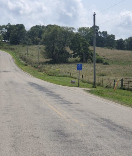 A rural road curves through a grassy landscape with trees and a signpost in the distance under a partly cloudy sky.