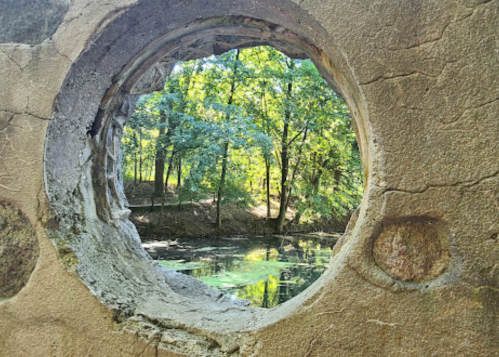 View through a circular stone opening, framed by trees and reflecting water in a serene natural setting.