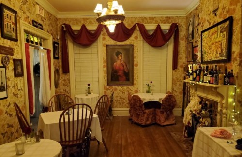 Cozy dining room with floral wallpaper, tables set for two, a large portrait, and a fireplace adorned with bottles.