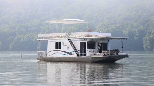 A houseboat floats on calm water, surrounded by lush green hills in a misty atmosphere.