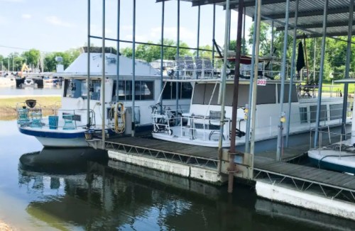 Two houseboats docked under a metal shelter at a marina, surrounded by calm water and greenery in the background.