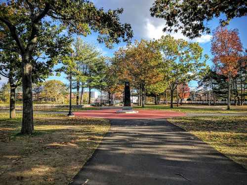 A pathway leads through a park with trees and colorful foliage, featuring a monument in the distance.