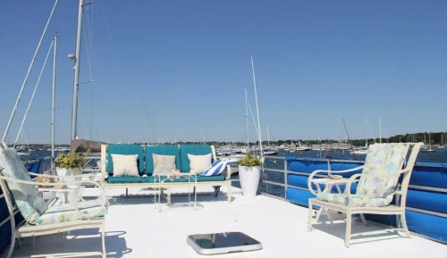 A sunny deck with turquoise cushions, white furniture, and a view of sailboats on the water under a clear blue sky.