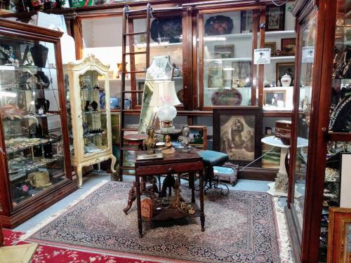 A cozy antique shop filled with glass display cases, vintage furniture, and decorative items on a patterned rug.