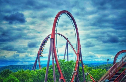 A roller coaster with loops against a cloudy sky, surrounded by green trees and distant hills.