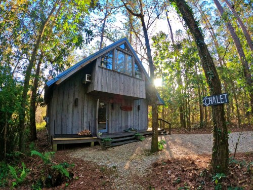A cozy wooden chalet nestled among tall trees, with a sign indicating "Chalet" and sunlight filtering through the leaves.