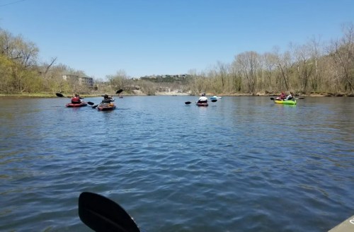 A serene river scene with several kayakers paddling on a sunny day, surrounded by trees and distant hills.