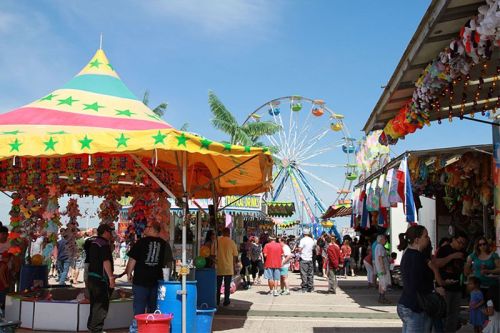A vibrant fair scene with colorful tents, a ferris wheel, and crowds enjoying games and attractions under a clear blue sky.