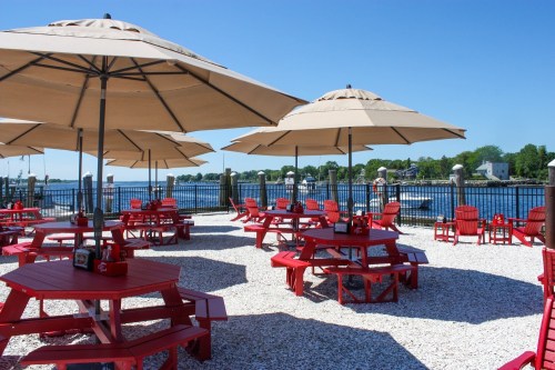 Outdoor dining area with red picnic tables and umbrellas by a waterfront, surrounded by a gravel surface and blue sky.
