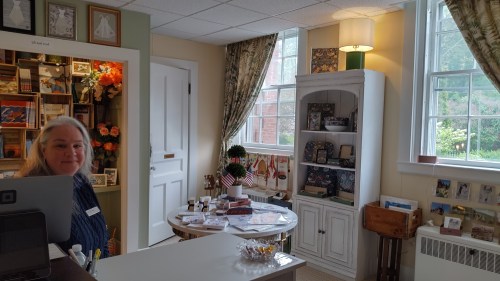 A woman stands at a counter in a cozy shop filled with crafts and decor, with windows letting in natural light.