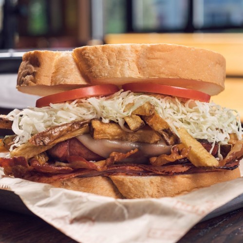 A hearty sandwich with layers of bacon, fried potatoes, coleslaw, and tomatoes between two slices of bread.