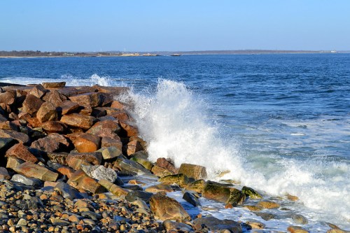Waves crashing against a rocky shoreline under a clear blue sky, with calm waters in the background.