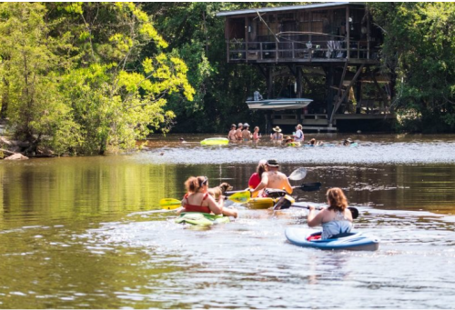 A group of people kayaking on a calm river, with a wooden cabin and others swimming in the background.