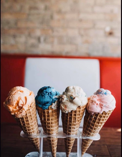Four ice cream cones in a display, featuring flavors of orange, blue, white, and pink against a brick wall backdrop.