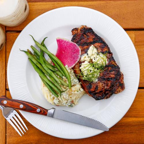 Grilled steak topped with butter, served with green beans, mashed potatoes, and a slice of radish on a white plate.