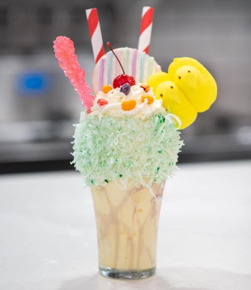 A colorful milkshake topped with whipped cream, coconut flakes, a cherry, candy, and decorative straws.