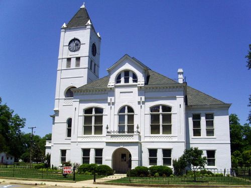 Historic white building with a clock tower, surrounded by greenery and a "Vote Here" sign in front. Clear blue sky.