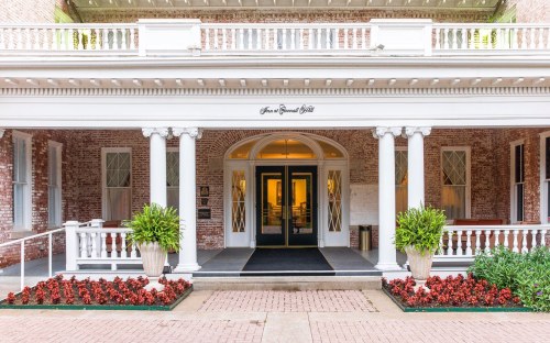 Entrance of a historic building with white columns, potted plants, and red flowers lining the walkway.