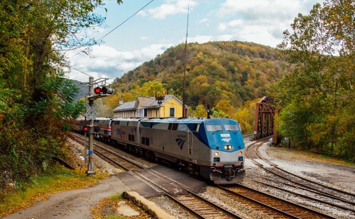 An Amtrak train approaches a crossing near a small station, surrounded by autumn foliage and rolling hills.