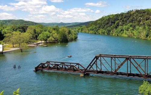A scenic river view with lush greenery, a partially submerged old railway bridge, and a small boat on the water.