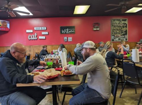 Two men eat at a busy restaurant with red walls, while other diners enjoy their meals in the background.