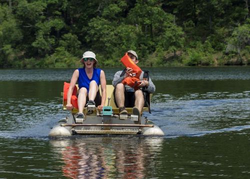 Two people sit on a small raft, enjoying a sunny day on a calm lake surrounded by greenery.