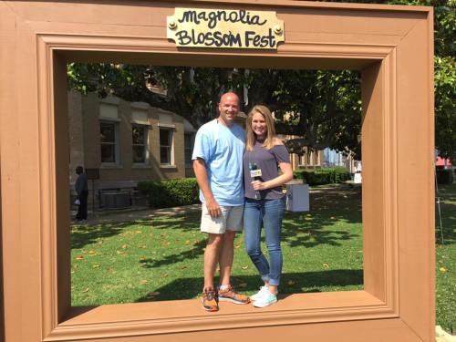 A couple poses together in a large frame at the Magnolia Blossom Fest, surrounded by greenery and buildings.
