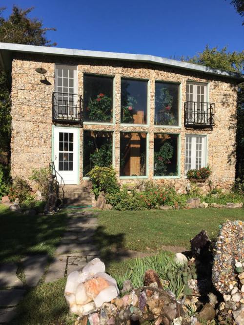 A unique stone house with large windows, surrounded by greenery and colorful flowers under a clear blue sky.