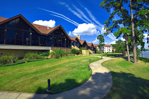 A scenic view of a building with a sloped roof, surrounded by green grass and trees under a blue sky with clouds.