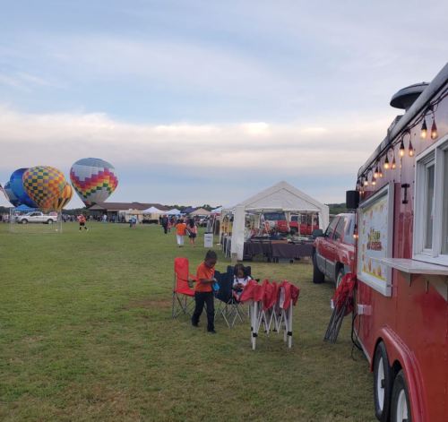 A food truck in a grassy field with colorful hot air balloons and people enjoying the event in the background.