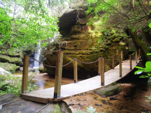 A wooden bridge with chains crosses a serene stream surrounded by lush greenery and moss-covered rocks.