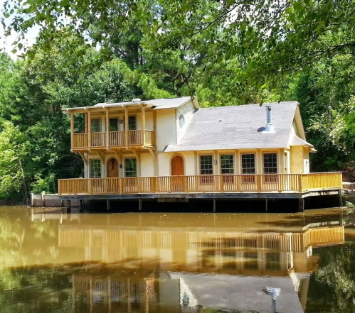 A charming wooden house on stilts by a calm lake, surrounded by lush green trees and reflecting in the water.