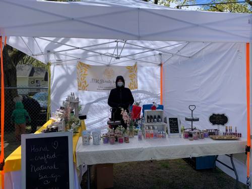 A vendor in a mask stands behind a table filled with beauty products at a market stall under a white tent.
