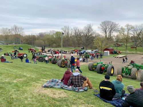 A crowd gathers on a grassy hill, watching tractors and activities at an outdoor event, with people sitting on blankets.