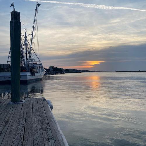 A serene sunset over calm waters, with boats docked at a wooden pier and a cloudy sky in the background.