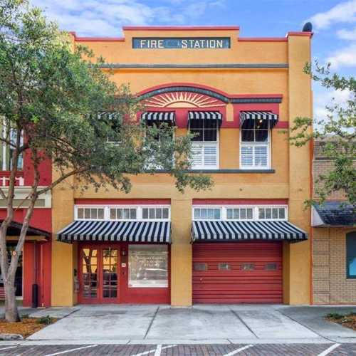 Historic fire station building with red and yellow facade, large windows, and an awning, surrounded by trees.