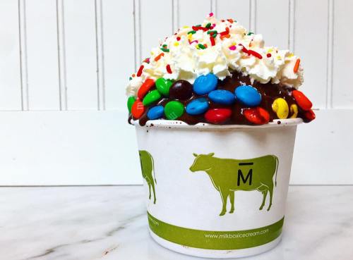 A cup of chocolate ice cream topped with whipped cream, colorful sprinkles, and M&M's, featuring a cow logo.