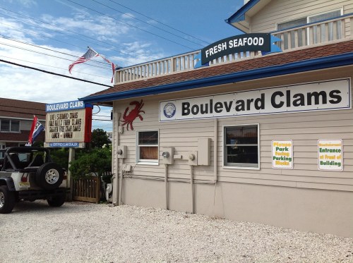 Exterior of Boulevard Clams seafood restaurant with signs and a parked vehicle in a gravel lot. American flags are visible.
