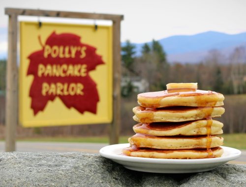 A stack of pancakes with syrup and butter in front of a sign for Polly's Pancake Parlor and a scenic background.
