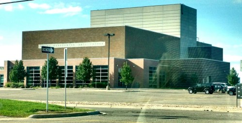 A modern performing arts center with large windows and a brick facade, surrounded by trees and a parking lot.